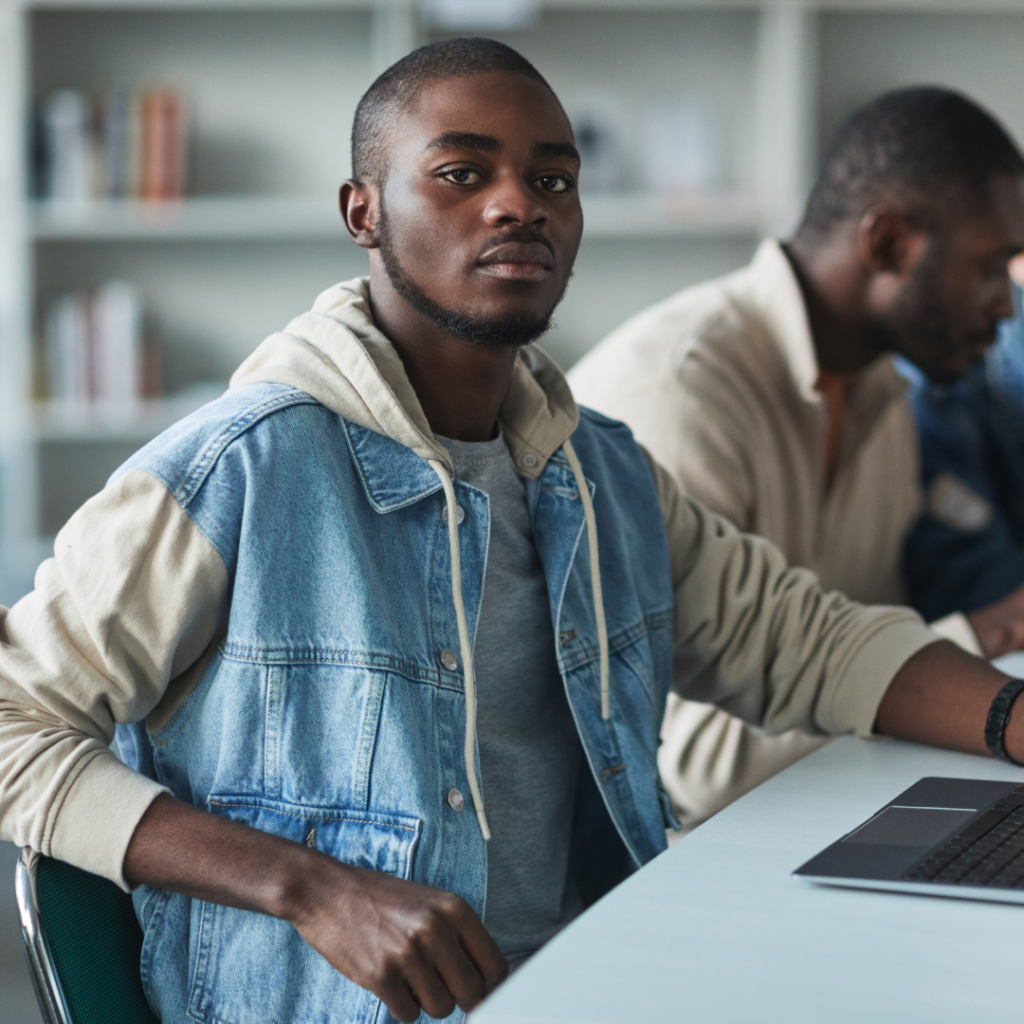 male student sitting wearing blue hoodie looking at the camera with another make student reading